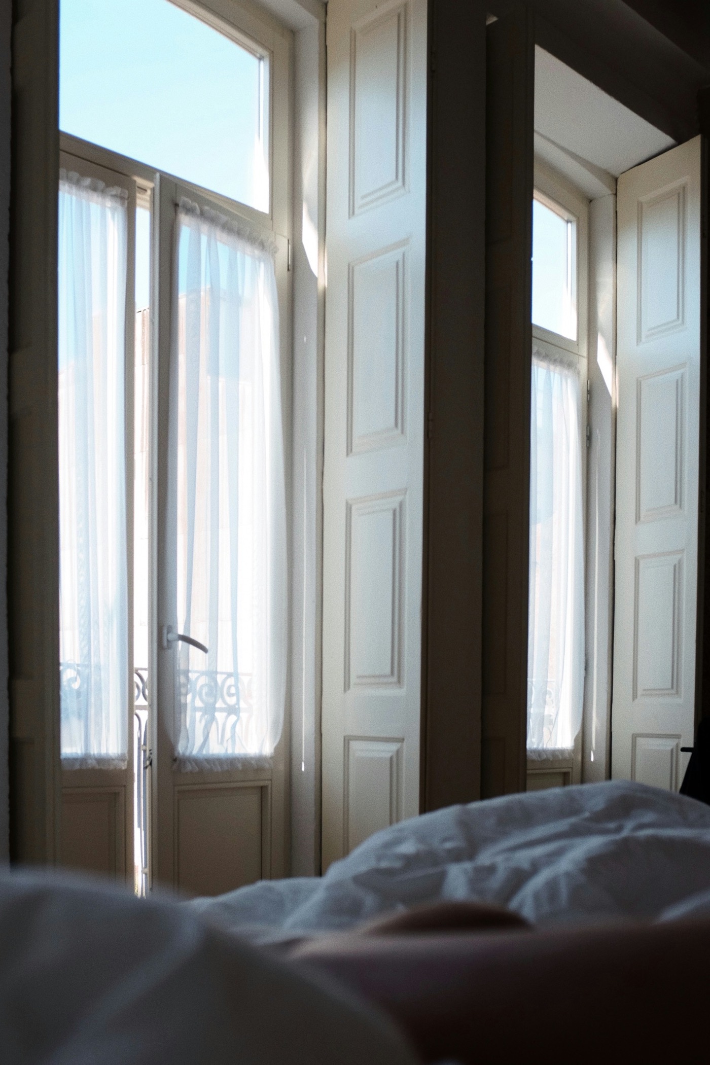 Grandes fenêtres à volets intérieurs et voilages baignés de lumière, couette au premier plan.