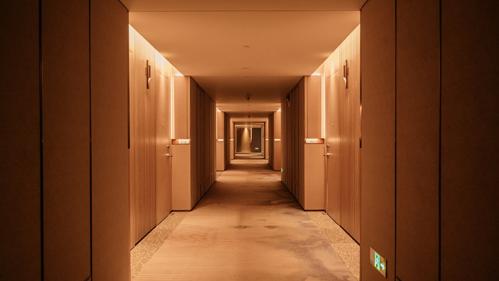 Couloir d’hôtel aux murs clairs, portes en bois et éclairage chaud.